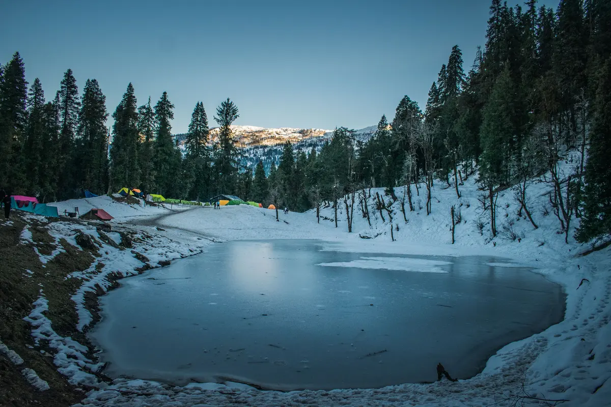 Frozen Juda Ka Taal lake ringed by pine forest and campsite tents