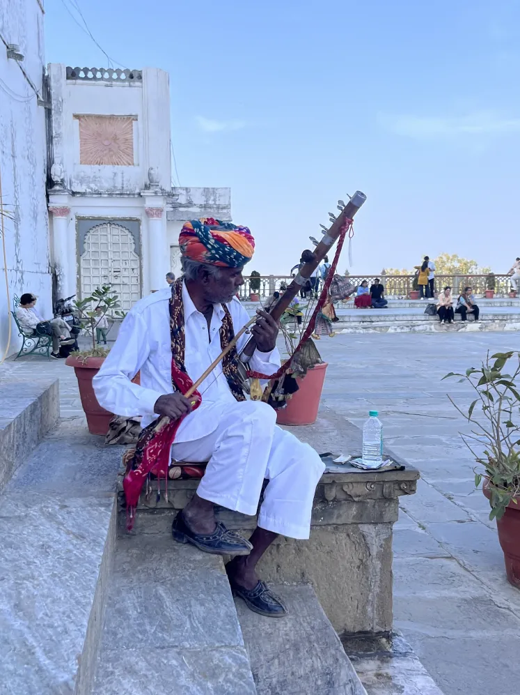 A Rajasthani folk musician in a bright turban playing a ravanhatha on the marble steps of a temple in Udaipur